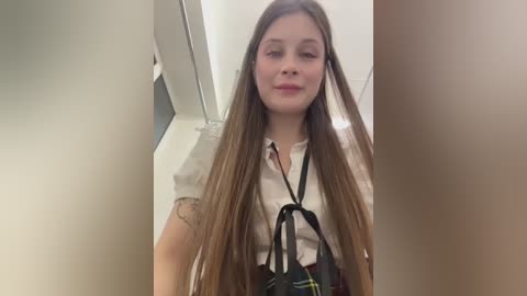 A video of a young woman with long, straight brown hair, wearing a white blouse and a black and yellow plaid skirt, standing in a brightly lit hallway.