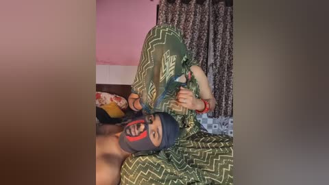 Video of a person wearing a black mask with red markings, lying on a bed with a green and white patterned sheet. The background features pink walls and floral-patterned curtains.