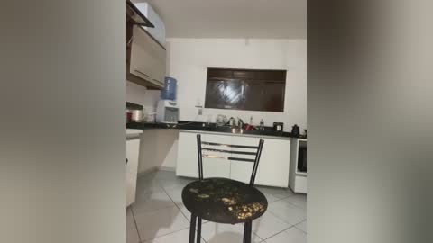 Video of a small, minimalist kitchen with white walls, black countertops, and a single wooden chair with a worn, rustic finish. The kitchen has basic appliances, including a water dispenser, and a window with a brown blind.