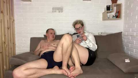 Video of a bald, shirtless man in navy briefs reclining on a beige sectional sofa, next to a woman with short blonde hair and glasses, wearing a white blouse and black shorts, holding a smartphone. They are in a white brick-walled room with wooden accents and shelves.