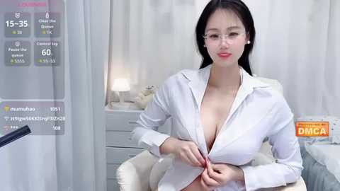 Video of a young East Asian woman with fair skin, long black hair, and large breasts, wearing a partially unbuttoned white lab coat, sitting in a medical examination room.