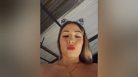 Video of a young woman with light skin, wearing cat ears and red lipstick, blowing a kiss, indoors with a corrugated metal ceiling and metal bars in the background.