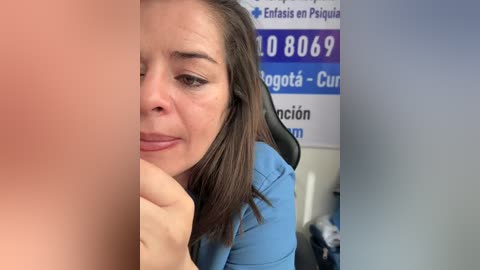 Video of a woman with light skin, shoulder-length brown hair, and light blue shirt, appearing distressed, in a clinical setting with phone numbers and signage in Spanish.