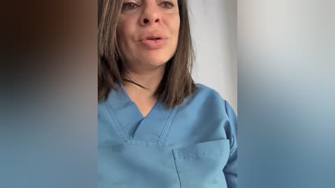 Video of a Caucasian woman with shoulder-length brown hair, wearing a light blue hospital scrub top, speaking in a medical setting with white walls and soft lighting.