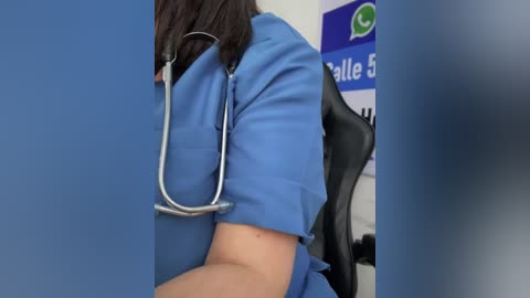 A video of a person in a blue medical uniform, with a stethoscope around their neck, sitting in an office chair. The background shows a white wall with a blue sign and a green logo.