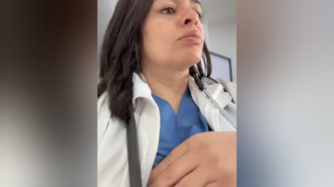 Video of a woman with light skin and dark hair, wearing a white lab coat over a blue scrub top, sitting with a thoughtful expression, holding her knee.