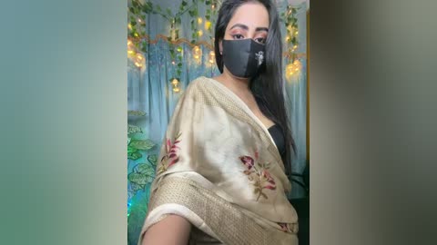 Video of a woman with long black hair, wearing a black mask, beige saree with floral patterns, and a black top. Background features green and blue curtains with fairy lights, creating a cozy ambiance.