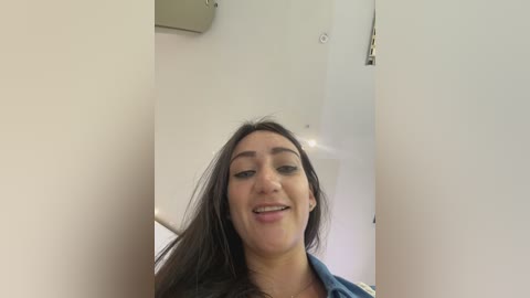 Video of a smiling Latina woman with long dark hair, wearing a blue shirt, standing in a modern white room with recessed lights.