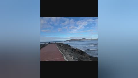 A video of a serene coastal walkway with a red brick path leading to rocky shorelines under a blue sky with scattered clouds.