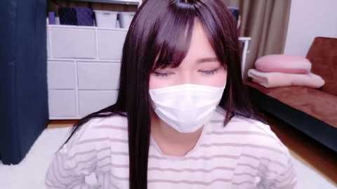 Video of a young Asian woman with long dark hair, wearing a white face mask, a striped shirt, and seated indoors, with a white dresser and a brown couch in the background.
