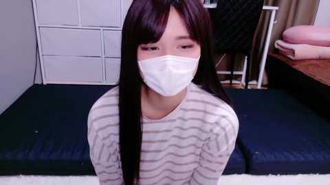 A video of an East Asian woman with long black hair, wearing a white surgical mask, striped shirt, and kneeling on a dark blue mat in a sparsely furnished room with white storage shelves and a bed.
