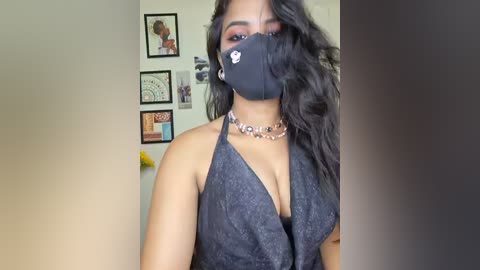 Video of a woman with medium-dark skin, wearing a black face mask, black halter top, and beaded necklace. Background features framed art and a yellow object.