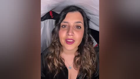 Video of a smiling woman with wavy dark hair, wearing dark lipstick and a black robe, lying on a bed with white sheets.