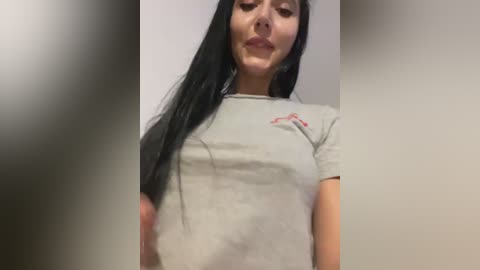 Video of a young woman with long, straight black hair and light skin, wearing a grey t-shirt. She has a neutral expression.