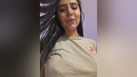 A video of a woman with long black hair, wearing a beige t-shirt, standing indoors. She has a neutral expression, and her hair is slightly disheveled. The background is blurred and indistinct.