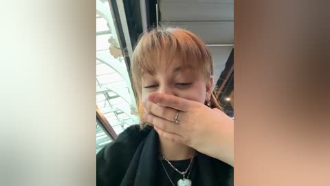 Video of a blonde woman with short hair, wearing a black robe, covering her mouth with her hand, standing in a room with green blinds and a white wall.