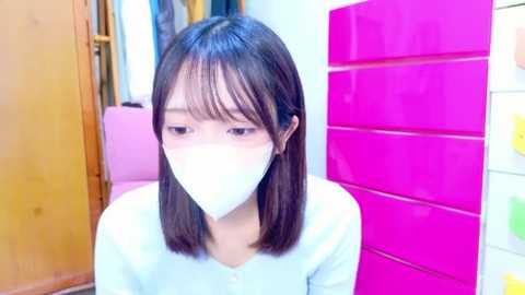 Video of an Asian woman with shoulder-length dark hair, wearing a white face mask, seated indoors next to a pink locker and wooden door.