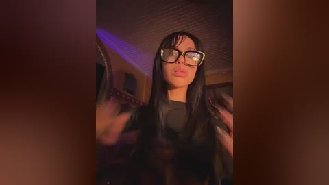 Media: A video of a woman with long black hair, wearing large black glasses, and a black top, standing in a dimly lit room with a purple glow.