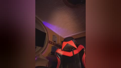 Video of a dimly lit room featuring a red and black gaming chair, with a large, textured, white ceiling fan and a hint of purple light.