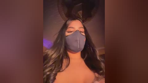 A video of a woman with long, black hair, wearing a gray mask and bunny ears, against a dark, warm-toned background.