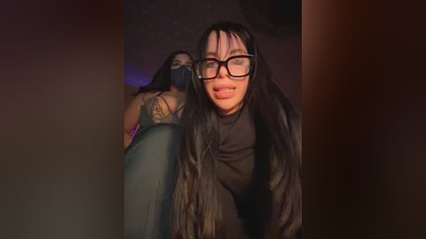 Media: Video of two women in dimly lit room: one with long black hair, glasses, dark top, smiling, and the other with dark hair, wearing a mask and pink top, in the background.