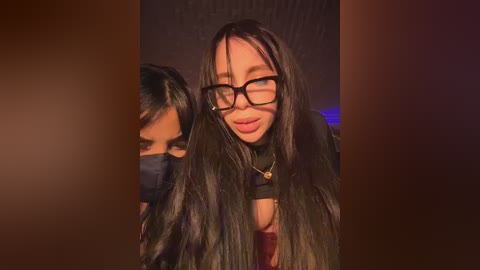 Video of a smiling young woman with long, straight black hair and black-framed glasses, wearing a black top, indoors against a dim, textured background.