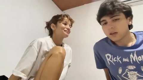 Video of two young men with light skin, one with short brown hair and the other with dark hair, wearing casual blue and white shirts, standing in a simple white room with a red ceiling corner.