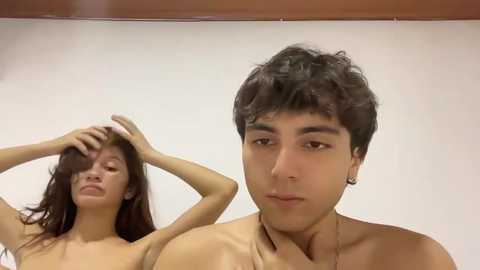 Video of a topless young woman with long brown hair, standing with her hands in her hair, looking distressed, and a young man with short dark hair, wearing a gold necklace, standing next to her, both in a plain white room.