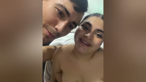 Video of a smiling, topless couple with medium skin tones, lying on a bed. The man, with short dark hair and a light beard, is on the left, and the woman, with dark hair tied back, is on the right.