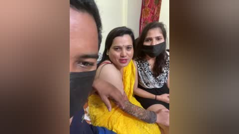 Video of three women in traditional attire, one with a yellow sari, another with a black mask, and a third with a black and white patterned blouse, sitting closely. The background features a red curtain.