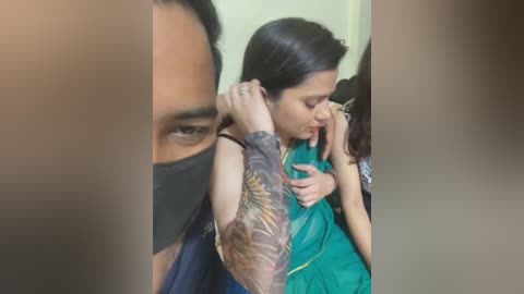 Video of a woman with long dark hair, wearing a green sari, comforting a crying child with a black mask, tattoos visible on her arm, in a dimly lit room.