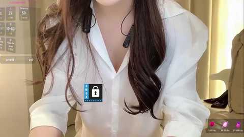 Video of a woman with long brown hair, wearing a white button-down shirt, headphones, and a \"Private\" badge, seated in a modern office with beige curtains and a digital clock.