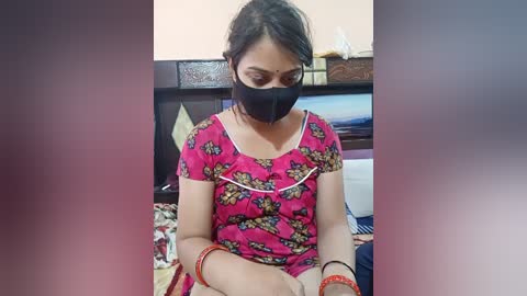 Media: Video of a South Asian woman with dark hair wearing a black face mask, pink floral dress, and red bracelets, sitting on a bed in a modest, cluttered room.