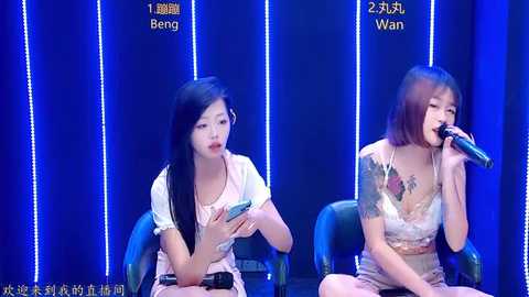 Video of two East Asian women with long black hair, wearing sleeveless dresses, seated at a microphone, singing against a blue, light-streaked backdrop.