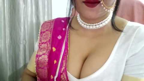 Video of a woman with medium skin tone, wearing a deep pink sari with gold embroidery, white blouse, gold choker, and nose ring, in a soft-lit room.