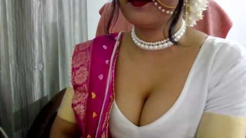 Video of a South Asian woman wearing a white blouse with a deep neckline, revealing ample cleavage, a pink sari with gold embroidery, and a pearl necklace.