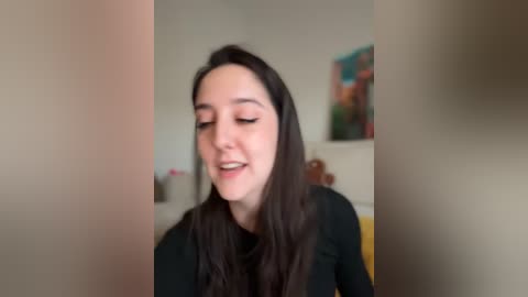 A video of a smiling woman with long dark hair, wearing a black top, standing in a softly lit room with a colorful painting on the wall.