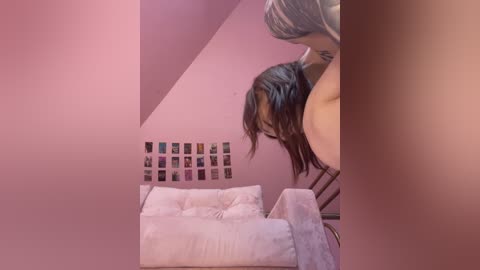 Video of a woman with long, dark hair and fair skin, leaning against a mirror in a pink room, featuring a white couch and a collage of photos on the wall.