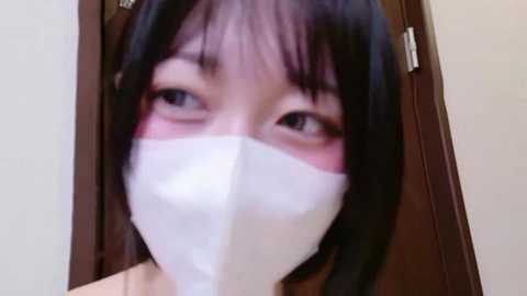 A close-up video of an East Asian woman with straight black hair and pale skin, wearing a white surgical mask, looking directly at the camera with a neutral expression. She is indoors with a wooden door in the background.