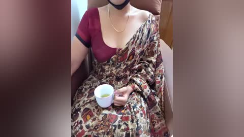 Video of a woman in a red blouse, black choli, and patterned sari, seated, holding a white cup, in a beige chair, indoors.