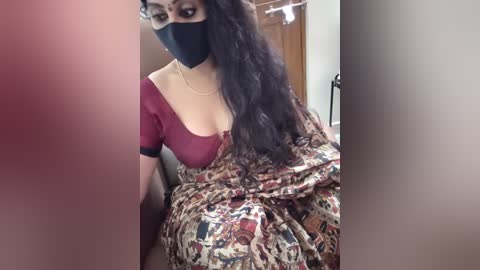 A video shows a woman with medium skin tone, wearing a maroon top with a sheer patterned sari, black face mask, and glasses, seated indoors with a blurred background.