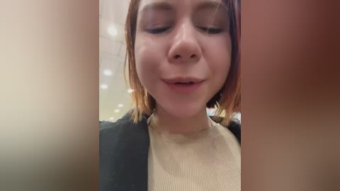 A close-up video of a young woman with fair skin and shoulder-length, light brown hair, wearing a beige sweater and black blazer. She has closed eyes and a slight smile.