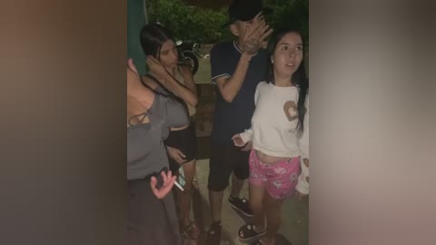 Video of four people in casual attire standing outdoors, one man with a cap covering his face, a young woman in a white long-sleeve shirt with a cartoon design and pink shorts.