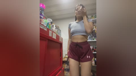 A video of a young woman in a supermarket, wearing a light blue crop top and maroon Under Armour shorts, standing in front of a red display rack.