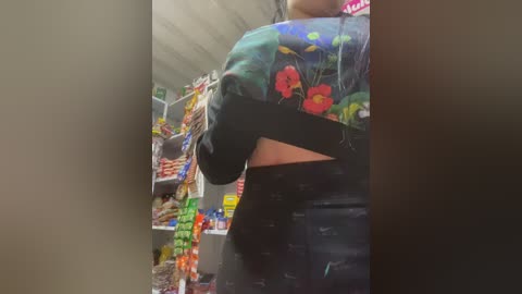 Video of a woman in a grocery store, wearing a colorful floral shirt and black leggings, standing in front of shelves filled with snacks and drinks.