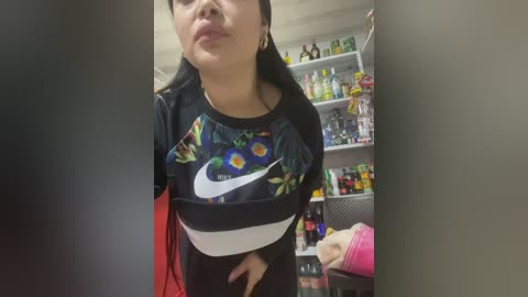 Video of a young woman with dark hair, wearing a Nike crop top with floral print, standing in a cluttered room filled with shelves of various items.