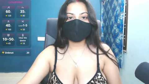 A video of a South Asian woman with long black hair, wearing a black face mask and leopard-print bra, seated indoors. Background features a blue wall and a colorful, floral-patterned curtain.