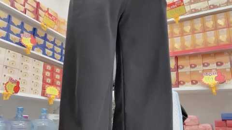 Video of a person's legs wearing black pants, standing in a brightly lit grocery store aisle with colorful, stacked shelves and promotional signs.