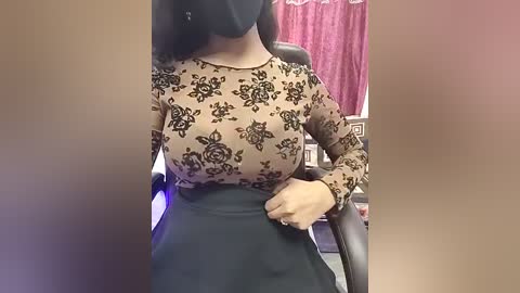 Video of a woman in an office, wearing a black mask, beige long-sleeve top with floral print, and high-waisted black skirt. She is adjusting her skirt while seated. Background includes a desk and pink curtain.