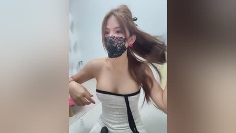 A video of an Asian woman with long brown hair, wearing a black lace mask, a gray strapless dress, and red earrings, standing in a bathroom.
